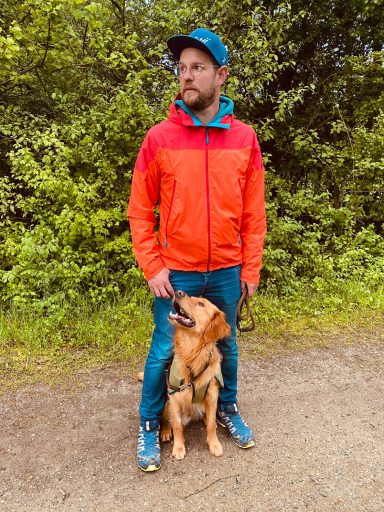 Mann in orange-roter Jacke steht neben einem Golden Retriever auf einem Weg.